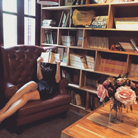 Femme lisant dans un fauteuil en cuir devant une bibliothèque en bois, ambiance chaleureuse avec bouquet de fleurs sur table en bois